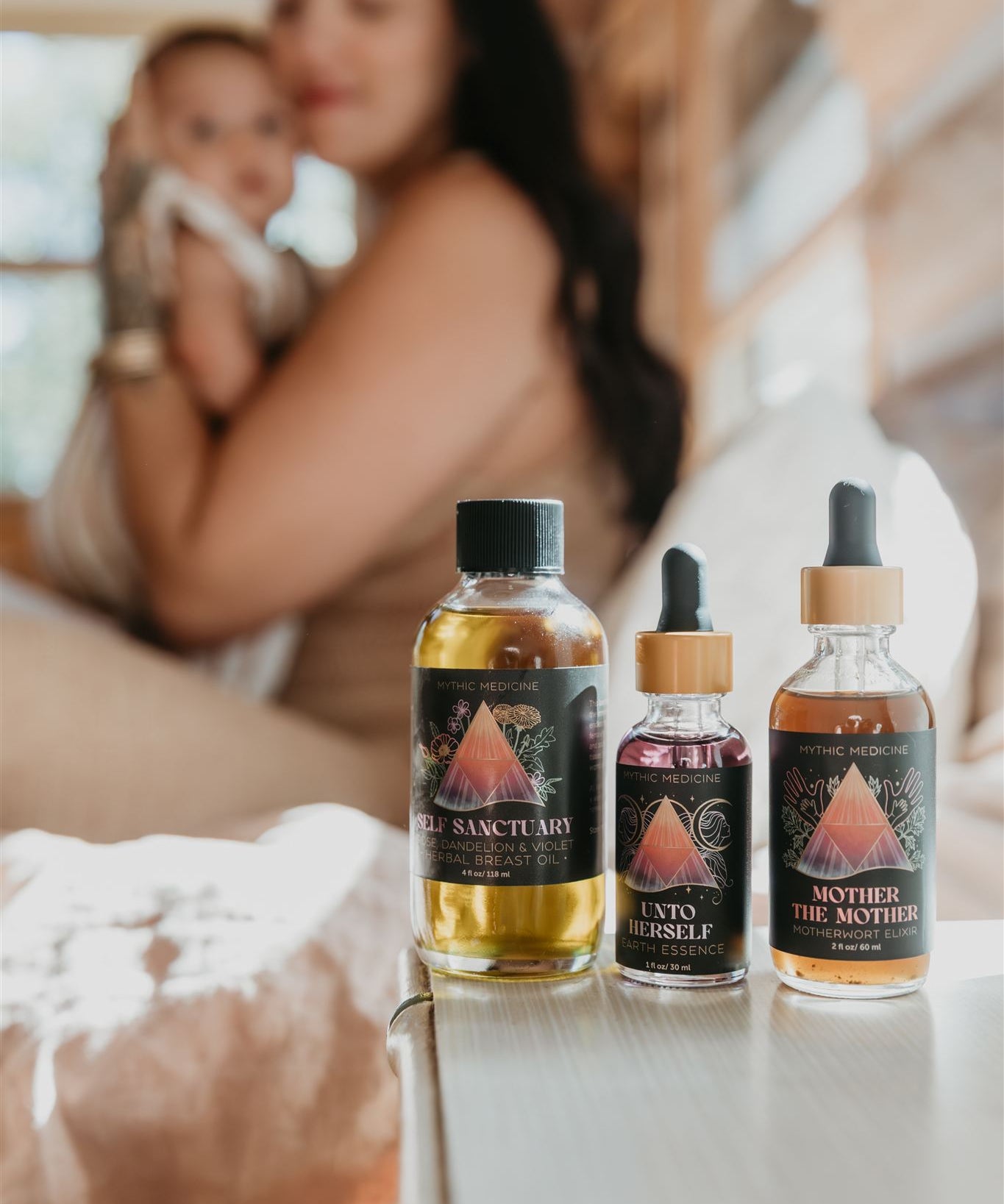 Three bottles of 'The Mother' Mythic Medicine on a surface with a blurred background of a woman holding a baby.