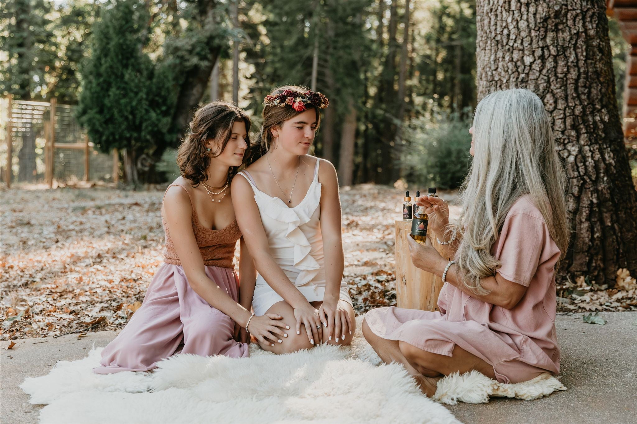 Two younger women sitting on a blanket outdoors near a tree and older woman holding Mythic Medicine bottle