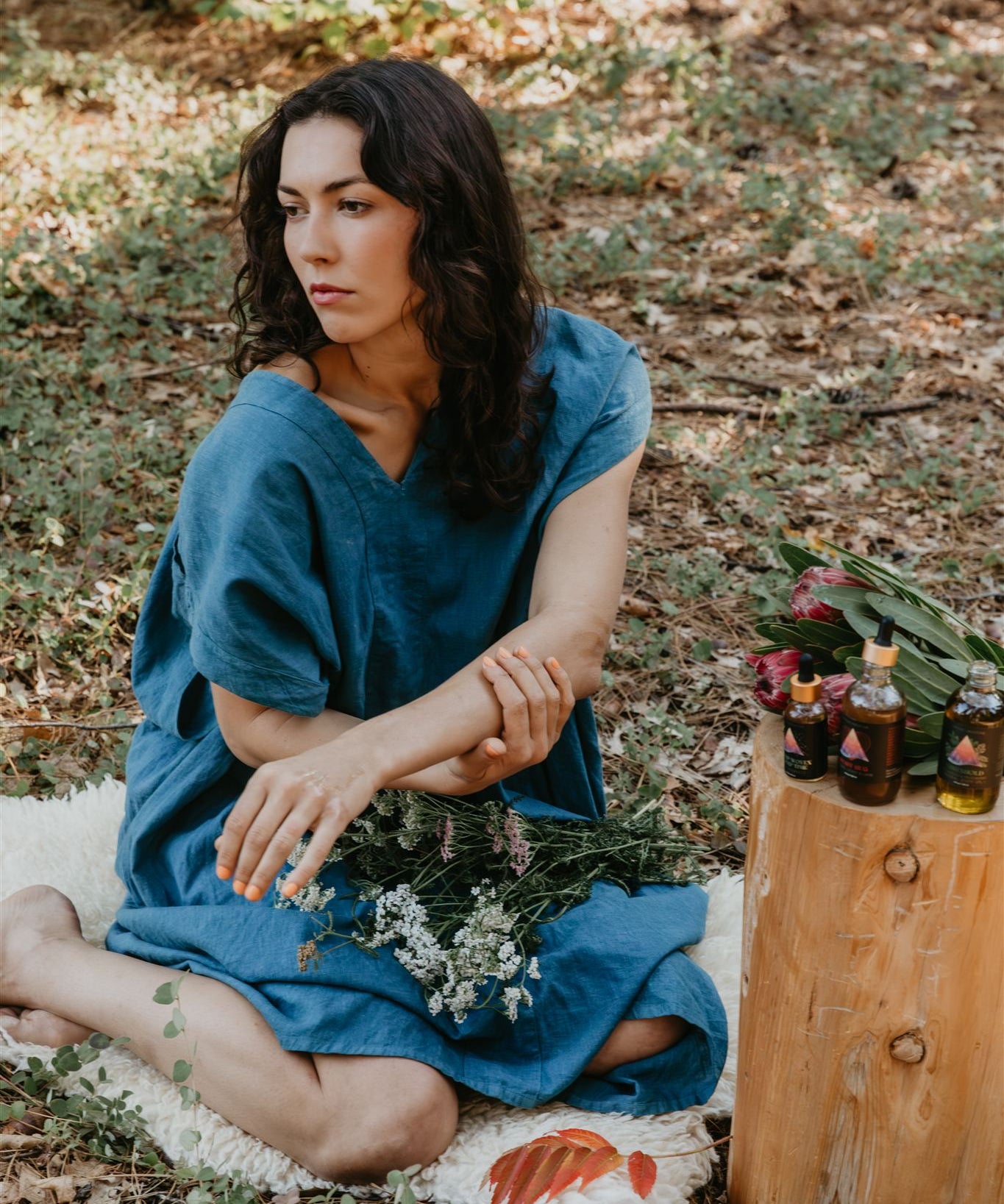 Woman in a blue dress sitting on a white rug with flowers in a forest setting.