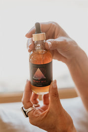 Hand holding a bottle of Mythic Medicine 'Heart Hug Herbal' tincture with a dropper against a neutral background