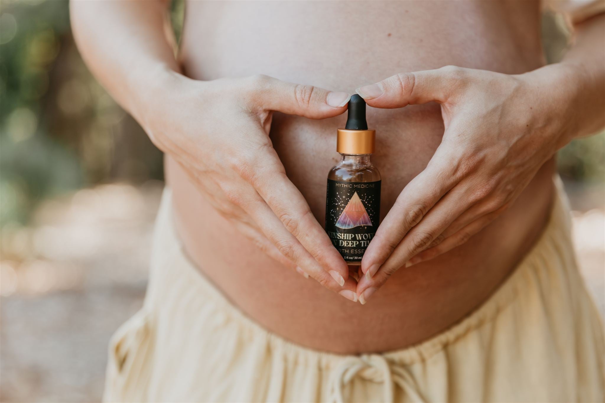 Woman holding a bottle labeled 'Mythic Medicine Kinship Oil' between their heart shaped hands with a blurred natural background