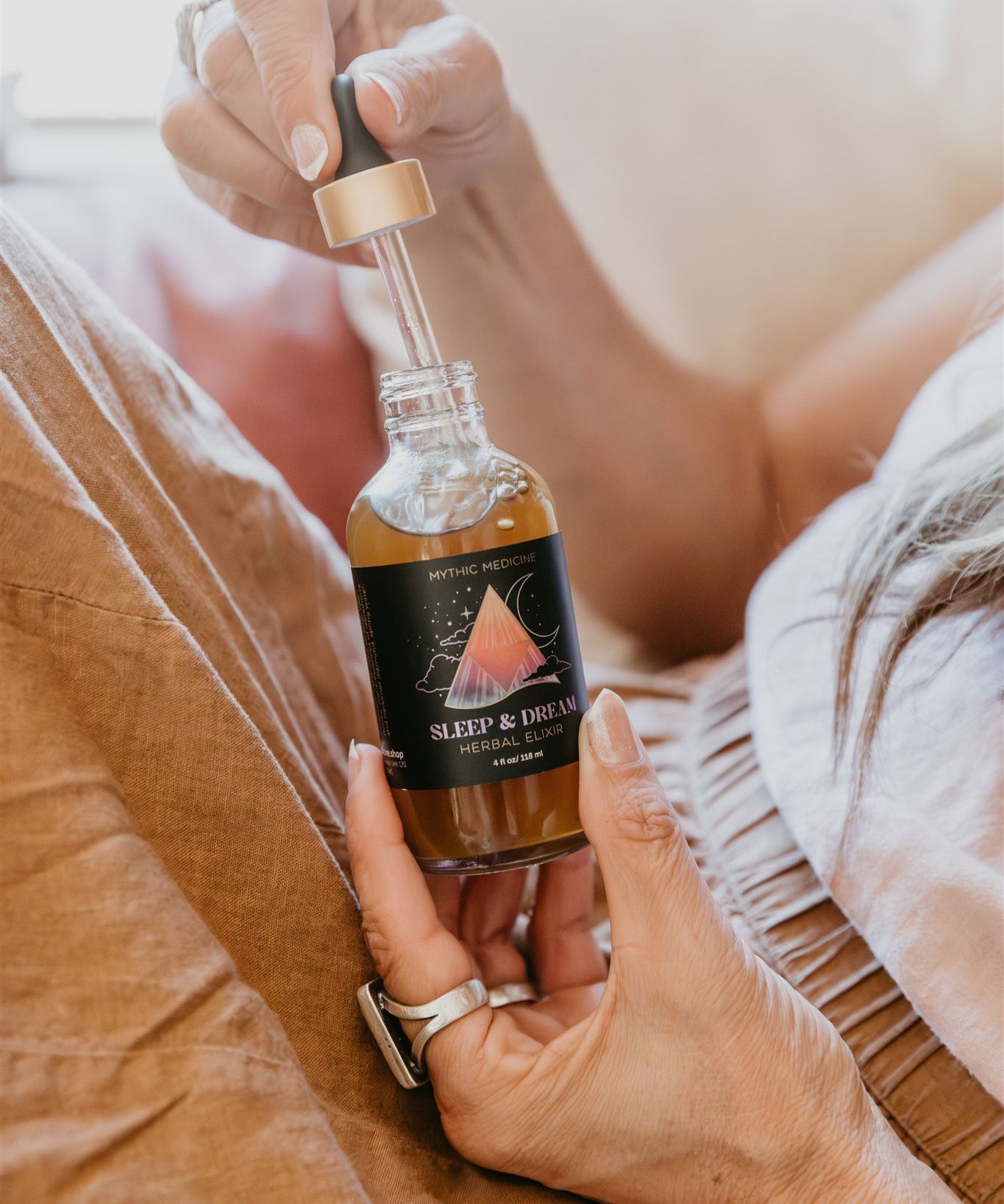 Person holding a bottle of Mythic Medicine 'Sleep & Dream' essential oil blend on a bed.