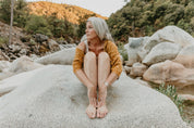 Woman with gray hair sitting on a rock by a stream with mountains in the background