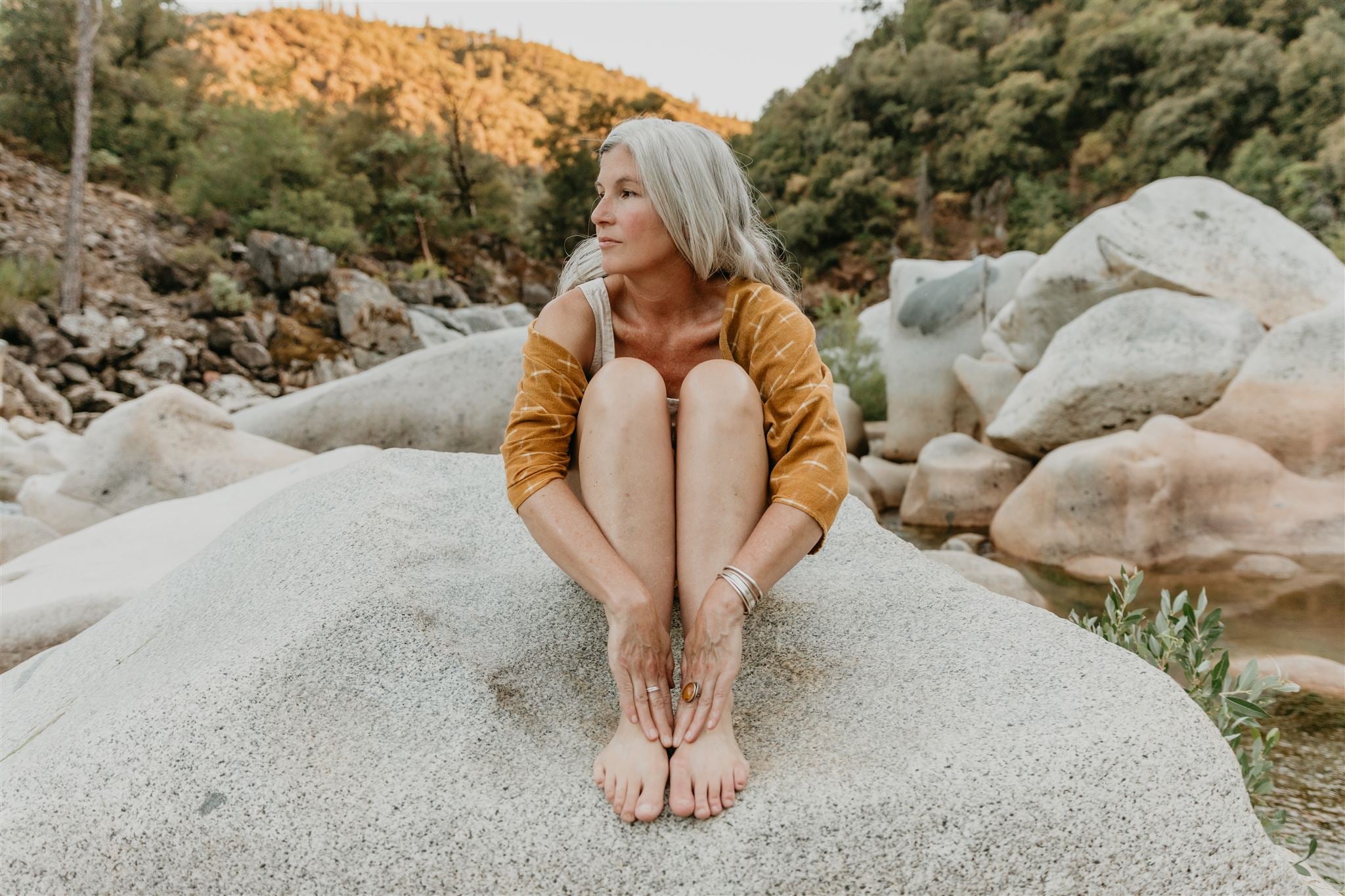 Woman with gray hair sitting on a rock by a stream with mountains in the background
