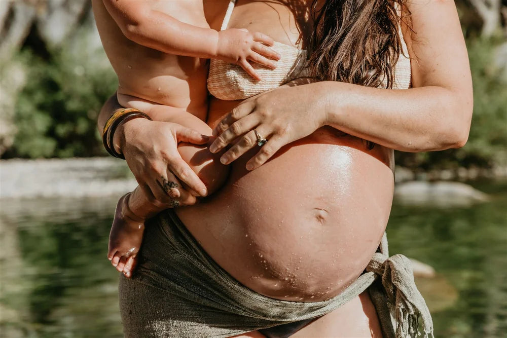 Pregnant bohemian woman with a child on her belly outdoors near water