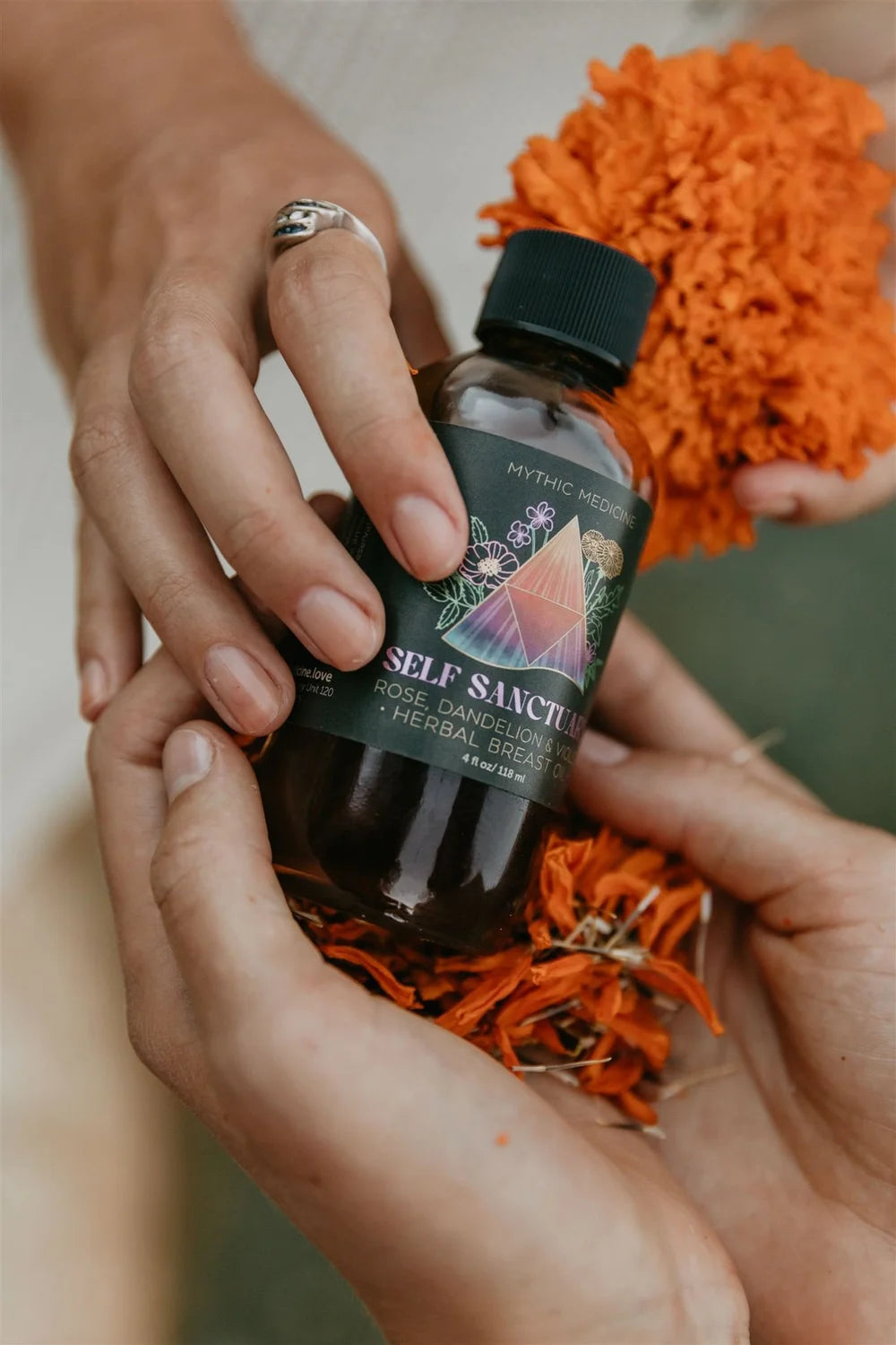 Bottle of 'Self Sanctify' with dried herbs held by hands against an orange textured background